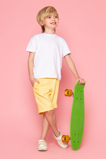 A front view cute smiling boy in white t-shirt and yellow jeans holding green skateboard on the pink space