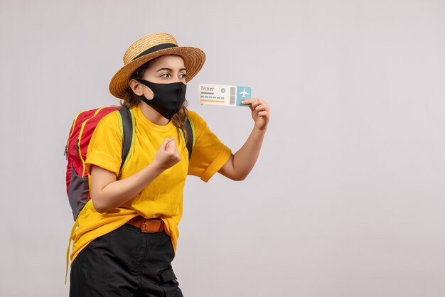 Front view of cute young traveller with backpack holding up travel ticket on grey isolated wall