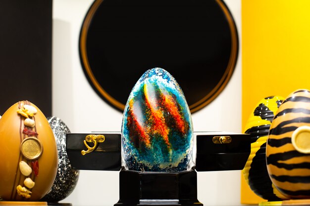 Front view decorated blue chocolate egg with red highlights on stand with other eggs