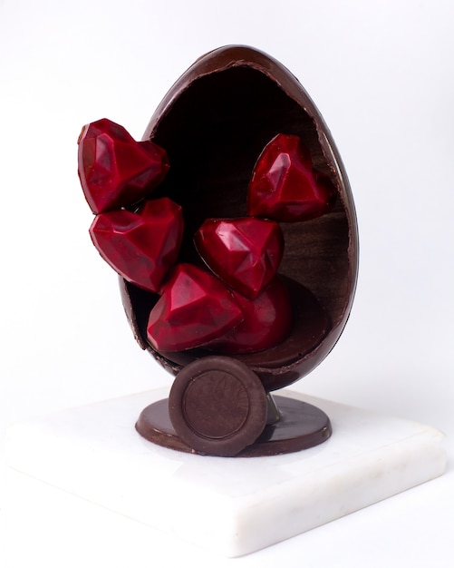 Вид спереди украшенное шоколадное яйцо с шоколадно-красными конфетами в форме сердца внутри на подставке