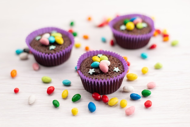 A front view delicious brownies inside purple forms with colorful candies on white, candy color sweets