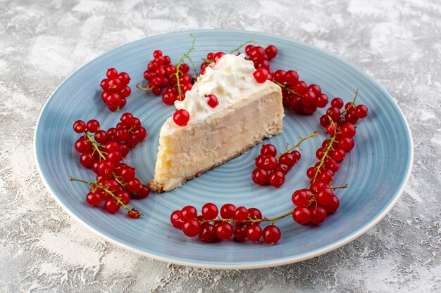 Вид спереди вкусного кусочка торта внутри синей круглой пластины с кремовой и красной клюквой на сером столе