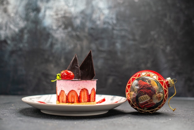 Free photo front view delicious cheesecake with strawberry and chocolate on oval plate and red xmas ball toy on dark