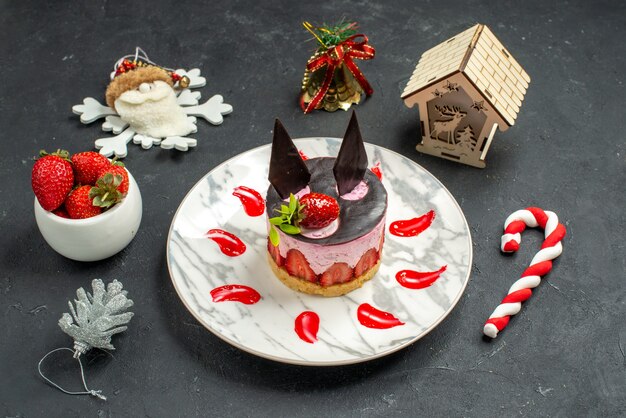 Вид спереди вкусный чизкейк с клубникой и шоколадом на тарелке, миске с игрушками из клубники на рождественской елке на темном фоне
