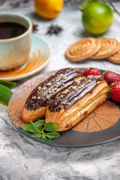 Вид спереди вкусные шоколадные эклеры с чаем на белом столе, печенье, десертный торт