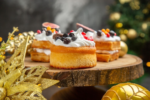 Front view delicious cream cakes around new year tree toys on dark desk dessert cake sweet photo cream