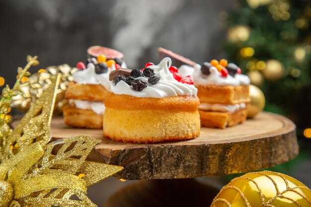 Front view delicious cream cakes around new year tree toys on dark desk dessert cake sweet photo cream