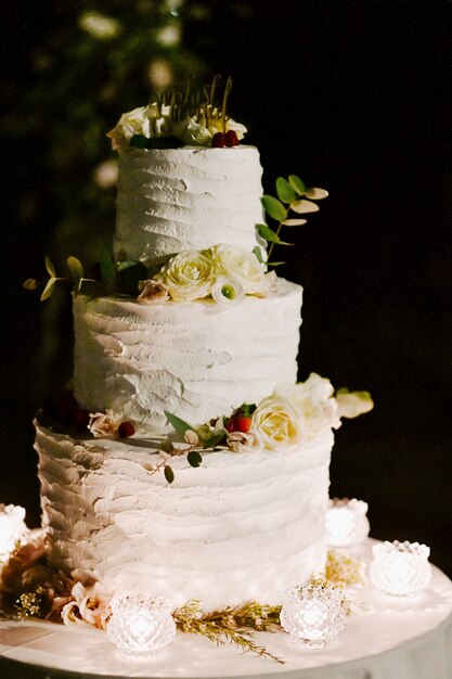 Front view of delicious creamy wedding cake decorated with eucalyptus and white roses on the table in the evening