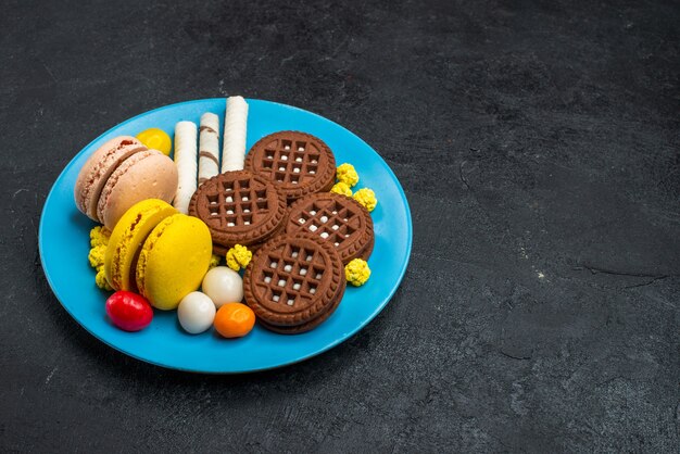 Вид спереди вкусные французские макароны с конфетами и шоколадным печеньем на серой поверхности бисквитного сахарного торта сладкого печенья