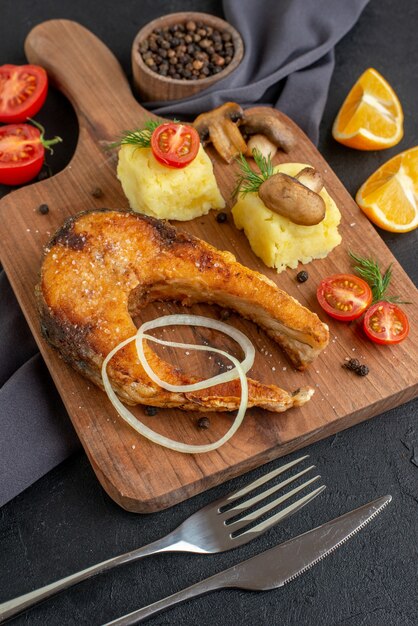 Front view of delicious fried fish meal with mushrooms vegetables cheese on wooden board lemon slices pepper on dark color towel cutlery set on black distressed surface