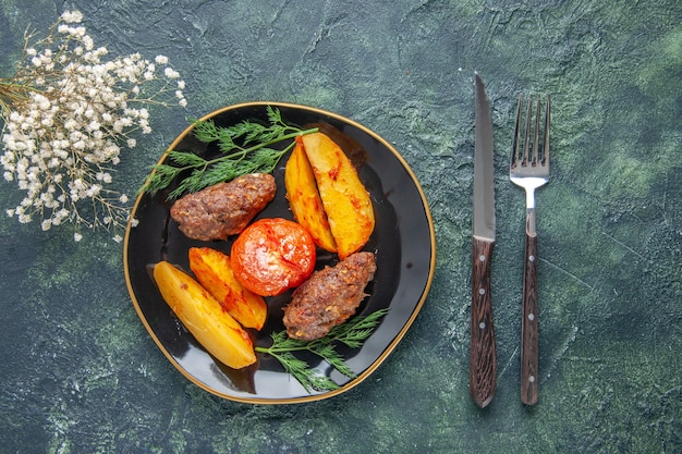Front view of delicious meat cutlets baked with potatoes and tomatoes on a black plate cutlery set white flowers on green black mixed colors background