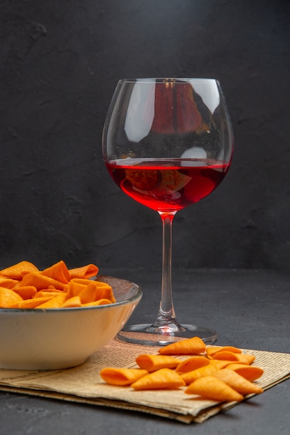 Front view of delicious potato chips inside and outside the bowl and red wine in a glass on an old newspaper