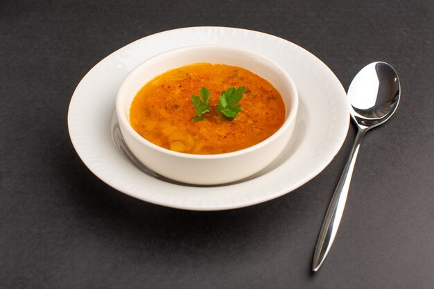 Front view of delicious soup inside plate with spoon on the dark desk