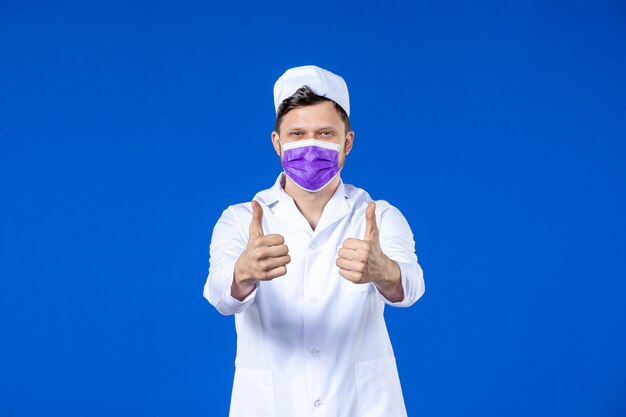 Front view of delighted male doctor in medical suit and purple mask on blue wall