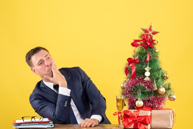 Vista frontale dell'uomo depresed che mette la mano sulla sua guancia che si siede al tavolo vicino all'albero di natale e presenta sul giallo