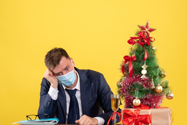 Front view of depressed business man sitting at the table near xmas tree and presents on yellow