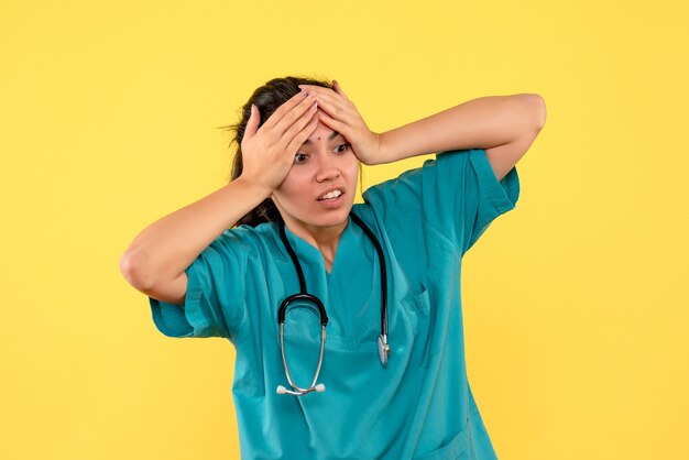 Вид спереди подавленная женщина-врач со стетоскопом, держащая голову, стоя на желтом фоне