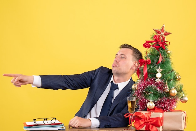 Front view of depressed man finger pointing something sitting at the table near xmas tree and presents on yellow