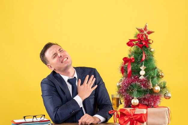 Front view of depressed man holding his chest with pain sitting at the table near xmas tree and gifts on yellow