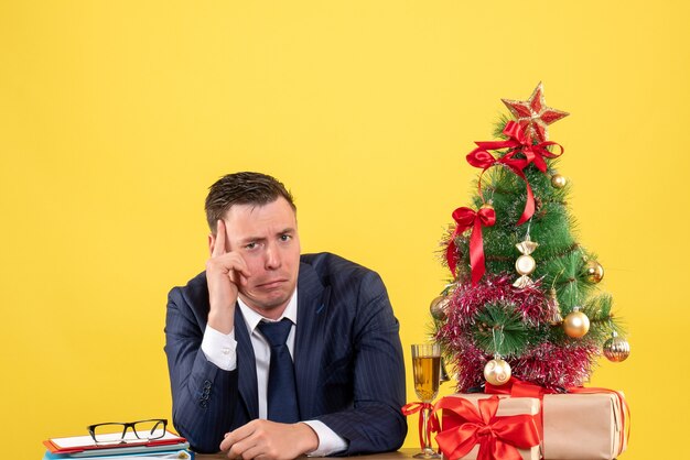 Front view of depressed man sitting at the table near xmas tree and gifts on yellow.