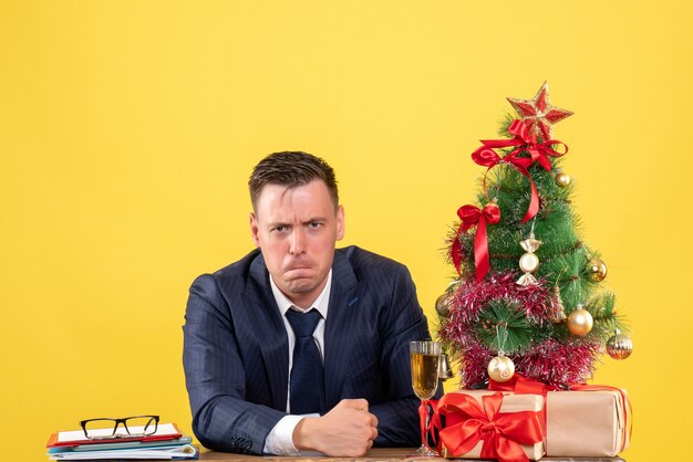 Front view of depressed man sitting at the table near xmas tree and presents on yellow.