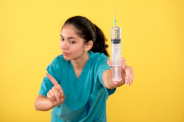 Front view determined woman doctor in uniform showing big syringe standing on yellow background