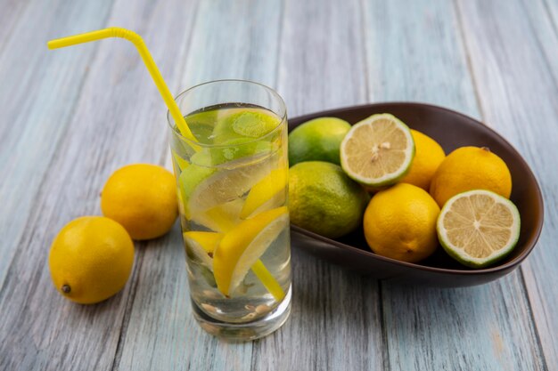 Front view detox water in a glass with a yellow straw and limes with lemons in a bowl on a gray background