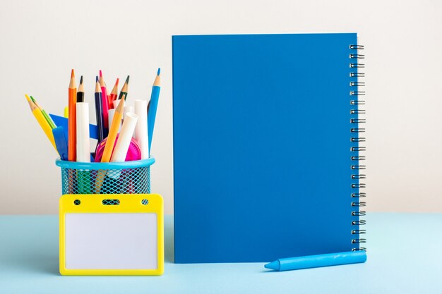 Front view different colorful pencils with blue copybook on blue desk