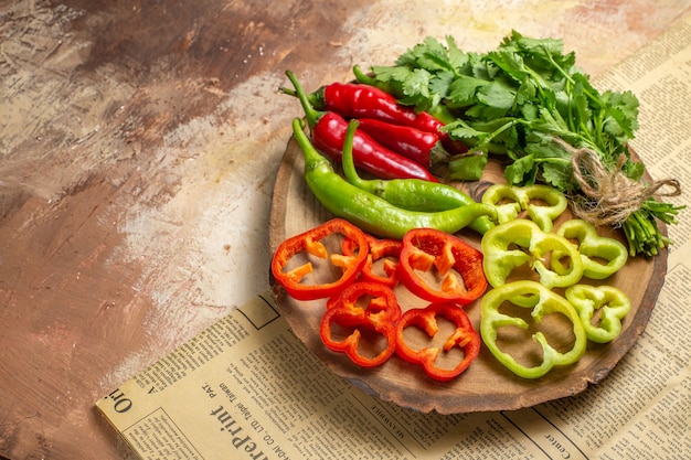 Front view different vegetables coriander hot peppers bell peppers cut into pieces on round tree wood board a newspaper on amber background with copy space