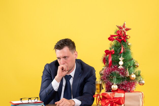 Front view of disappointed man sitting at the table near xmas tree and gifts on yellow.