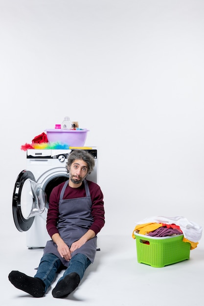 Front view dispirited housekeeper man sitting near laundry basket on white background