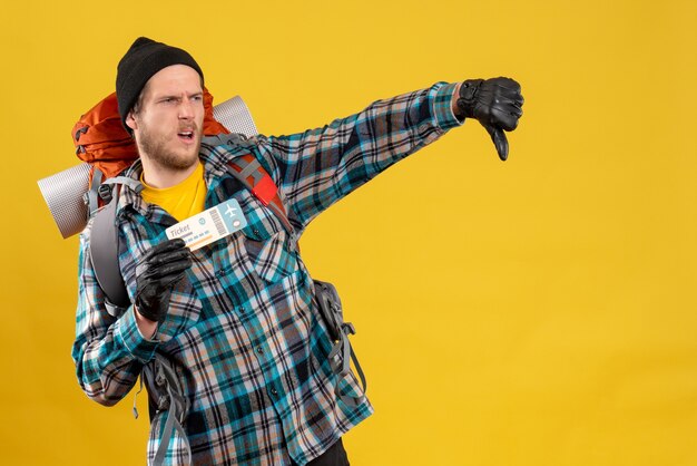 Front view of dissatisfied young backpacker with black hat holding plane ticket giving thumb down