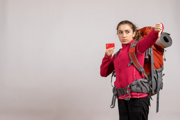 Вид спереди недовольного молодого путешественника с большим рюкзаком, держащего карточку, делающую большой палец вниз знак