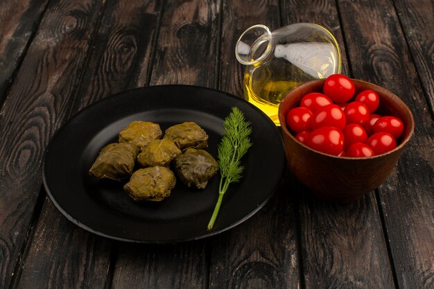 вид спереди долма внутри черной тарелки вместе со свежими красными помидорами черри и оливковым маслом на коричневом деревянном деревенском полу