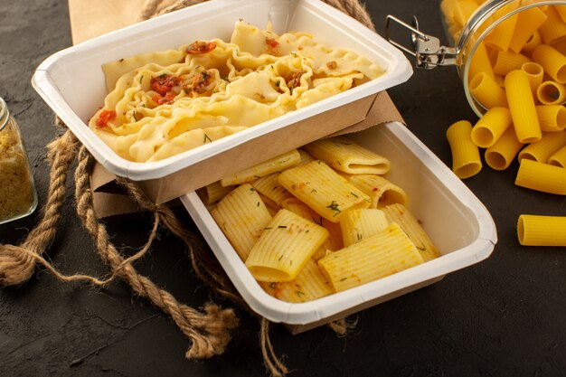 A front view dough meal with dried green herbs inside white bowls and package along with raw italian pasta on dark