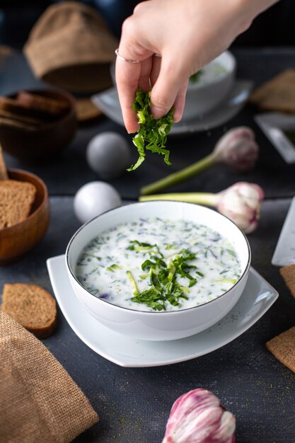 A front view dovga dish with dried mint getting poured with greens inside white plate along with bread loafs eggs flowers on the table soup liquid hot on the grey desk