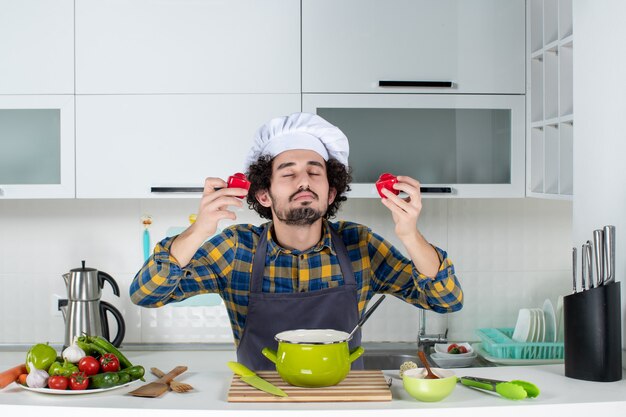 Front view of dreamy male chef with fresh vegetables holding red peppers in the white kitchen