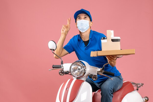 Front view of dreamy male delivery person in mask wearing hat sitting on scooter delivering orders on pastel peach background