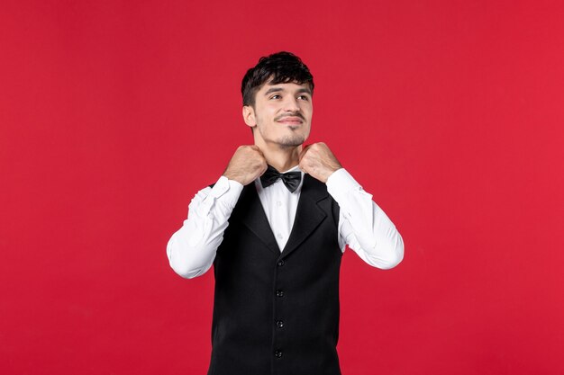 Front view of dreamy male waiter in a uniform touching butterfly on neck on red background