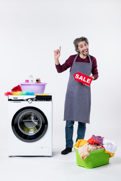 Front view elated guy in apron holding up sale sign standing near washing machine on white background