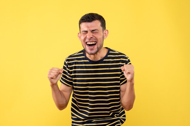 Front view elated handsome man in black and white striped t-shirt showing winning gesture on yellow isolated background