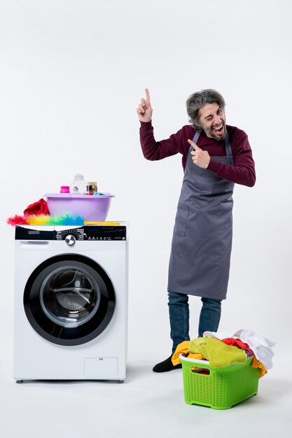 Front view elated housekeeper man pointing with finger up standing near washer laundry basket on white background