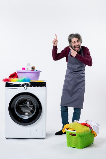 Front view elated housekeeper man standing near washer laundry basket on white background