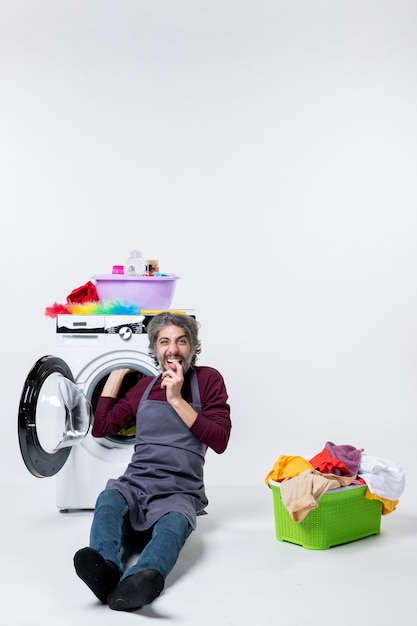 Front view elated male housekeeper sitting in front of washer green laundry basket on white isolated background