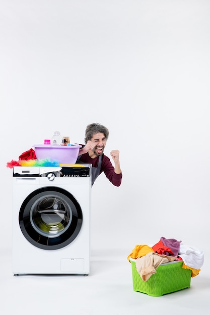 Front view elated man in apron sitting behind washing machine laundry basket on white background