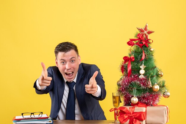 Front view of elated man finger pointing camera sitting at the table near xmas tree and presents on yellow