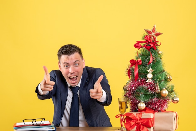 Вид спереди возбужденного человека, указывающего пальцем на камеру, сидящего за столом возле рождественской елки и подарков на желтом