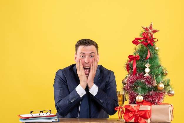 Вид спереди восторженного человека, кладущего руки на щеку, сидящего за столом возле рождественской елки и подарков на желтом