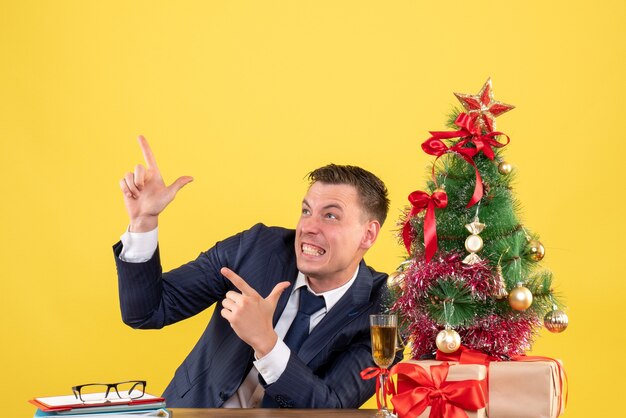 Front view of elated man sitting at the table finger pointing something near xmas tree and gifts on yellow