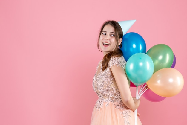 Front view elated party girl with party cap holding balloons behind her back